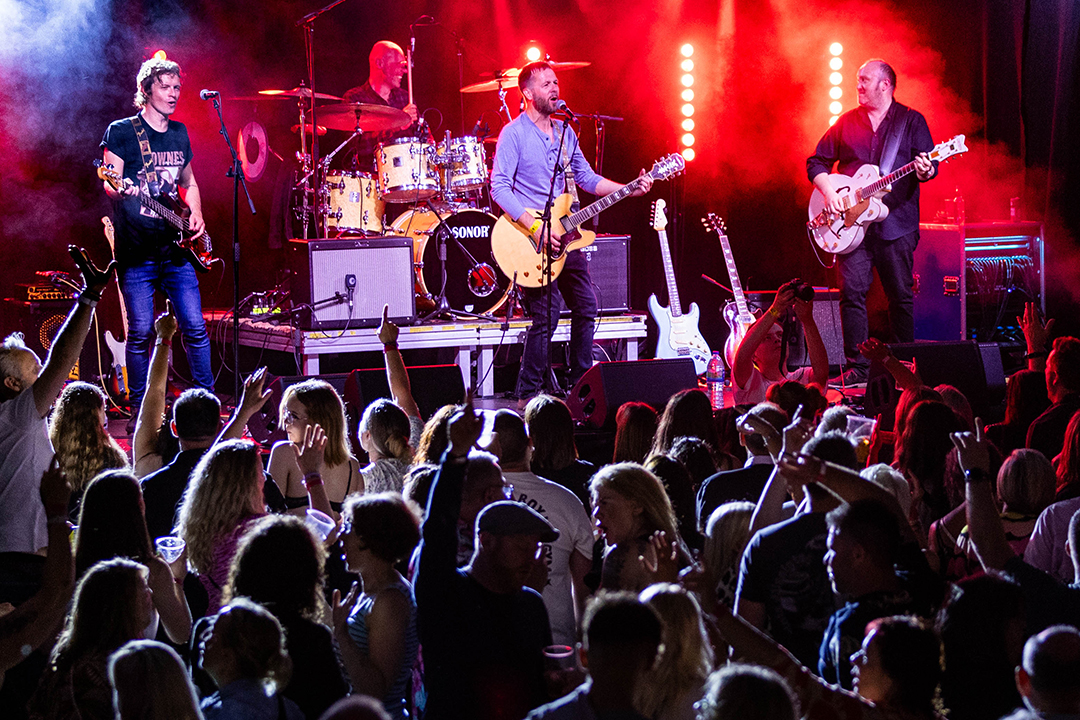 Four members of a band performing on stage in front of a big audience of people standing up and dancing